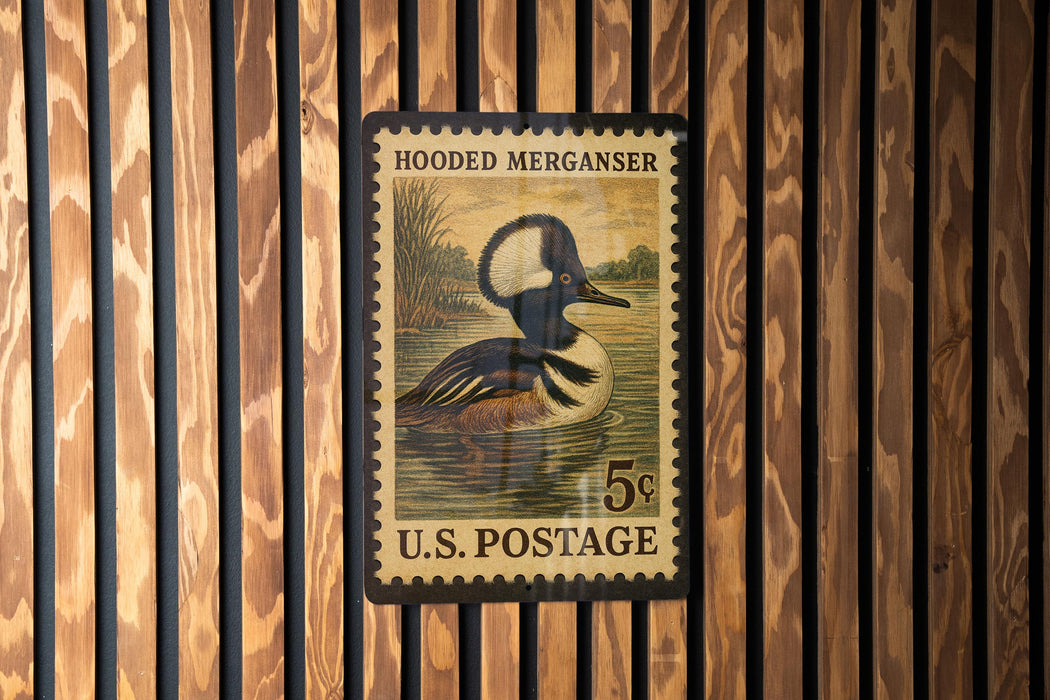 A U.S. postage stamp featuring a hooded merganser duck, mounted on a wooden wall.