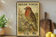 A framed illustration of a red bird, likely a finch, perched on a tree branch, with a U.S. 8¢ postage stamp in the background.