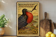a large, colorful postage stamp depicting a magnificently frigatebird, with the text "MAGNIFICENT FRIGATEBIRD" and "AIGATUGUA 20" printed on it. The stamp is displayed on a shelf next to a wooden cutting board and a potted plant.