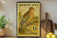 A vintage postage stamp featuring a mourning dove perched on a branch, with the text "MOURNING DOVE" and "U.S. POSTAGE" printed on it. The stamp is displayed on a shelf next to a wooden cutting board and a potted plant.