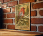 A wooden frame with a framed illustration of a red bird, likely a pine grosbeak, on a brick wall.