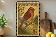 A vintage-style postage stamp featuring a red cardinal perched on a branch, with a potted plant and wooden cutting board in the background.