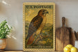 A framed vintage U.S. postage stamp depicting a red-tailed hawk is displayed on a shelf next to a wooden cutting board and a potted plant.