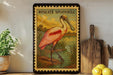 A framed illustration of a pink spoonbill bird is displayed on a shelf, accompanied by a wooden cutting board, a potted plant, and some lemons.