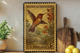 A vintage postage stamp featuring a hummingbird, with the text "Rufous Hummingbird" and "U.S. Postage 3¢" printed on it. The stamp is displayed on a shelf next to a wooden cutting board and a potted plant.