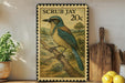 A framed postage stamp featuring a blue and green bird perched on a branch, with a rural landscape in the background.