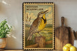 A framed illustration of a yellow bird, likely a meadowlark, perched on a wooden post against a natural landscape.