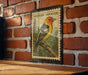 A metal stamp with a colorful illustration of a bird, likely a Western Tanager, perched on a branch against a brick wall.