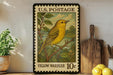 A vintage U.S. postage stamp featuring a yellow warbler, with a price of 10 cents, is displayed on a shelf next to a wooden cutting board and some kitchen utensils.