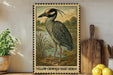 A framed illustration of a yellow-crowned night heron is displayed on a shelf, accompanied by a wooden cutting board, a potted plant, and some lemons.