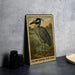 A framed illustration of a yellow-crowned night heron is displayed on a wall, with a plate and a stack of black camera lenses to the left.