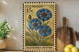 A vintage U.S. postage stamp featuring a blue flower design, placed on a wooden surface next to a wooden cutting board and a potted plant.