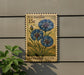 A U.S. postage stamp featuring a blue flower is displayed on a wall, with a plant to the left.