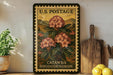 A framed vintage U.S. postage stamp depicting a large pink flower, with the text "U.S. POSTAGE" and "CATAWBA RHODODENDRON" printed on it. The stamp is displayed on a shelf next to a wooden cutting board and a potted plant.