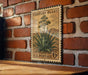 A vintage-style metal postage stamp featuring an illustration of an agave plant, mounted on a brick wall.