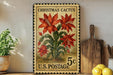 A vintage Christmas cactus postage stamp from the U.S. Postal Service, featuring a large red flower surrounded by green leaves, displayed on a shelf next to a wooden cutting board and a potted plant.