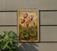 a framed illustration of pink flowers on a wall, with a plant to the left of the frame.