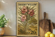 A framed vintage postage stamp featuring a floral illustration of pink flowers, placed on a wooden shelf next to a wooden cutting board and a potted plant.