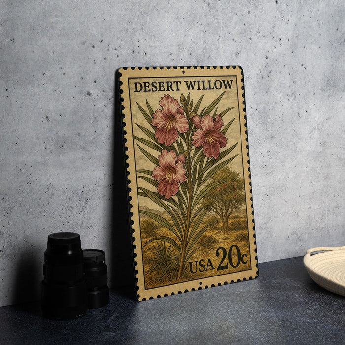 A vintage Desert Willow postage stamp from the United States, featuring a floral illustration of pink flowers and green leaves, is displayed on a gray countertop next to a stack of black camera lenses.