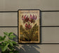 a framed illustration of a purple flower on a wall, with a plant to the left of the frame.
