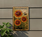 a framed illustration of three large sunflowers on a wall, with a plant to the left of the frame.