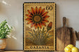 a framed illustration of a flower, specifically a Gazania, on a shelf. The frame has a stamp-like appearance and the word "Gazania" is written on it. There are also some kitchen utensils, such as a wooden cutting board and a knife, placed next to the frame.