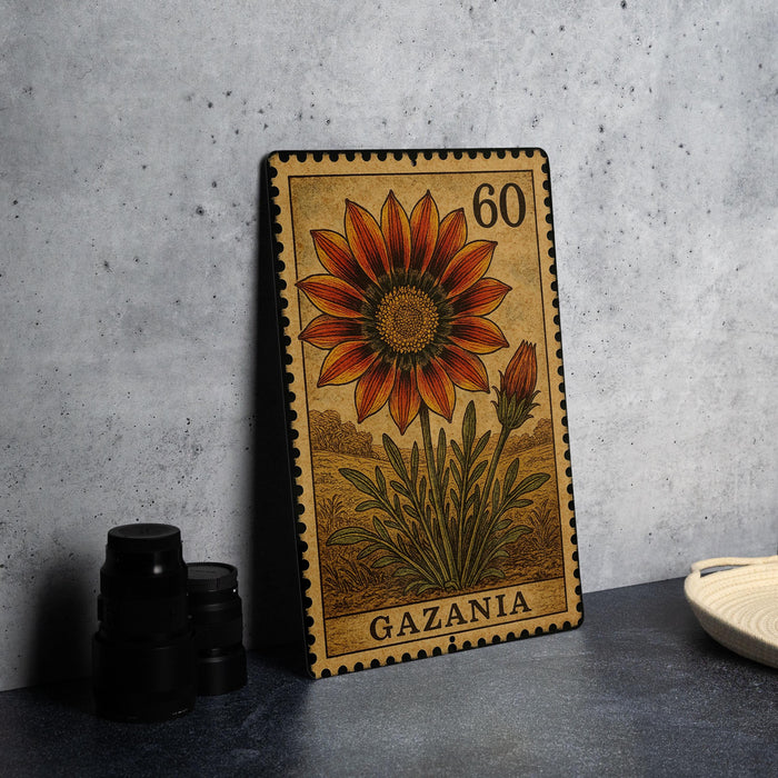A metal stamp from Gazania featuring a large orange flower with a yellow center, placed on a gray countertop next to some black camera lenses.