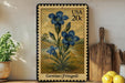 A framed postage stamp featuring a blue iris flower is displayed on a shelf next to a wooden cutting board and a potted plant.
