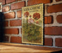 A vintage-style poster of geranium flowers is displayed on a brick wall, with a wooden table below it.