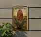 A postage stamp featuring a large orange flower with green leaves, mounted on a wall next to a plant.