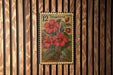 A vintage postage stamp featuring a floral design with red flowers and green leaves, mounted on a wooden wall.
