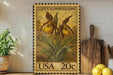 A framed stamp depicting a yellow orchid plant, with the text "LADY'S SLIPPER YELLOW" and the stamp value "20c" printed on it. The stamp is placed on a wooden surface, with a potted plant, a wooden cutting board, and some lemons nearby.