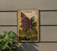 A framed illustration of a purple lilac flower is displayed on a wall, with a plant visible to the left.