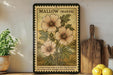 A vintage postage stamp of a marshmallow plant, labeled as 'Mallow (Marsh)', is displayed on a wooden shelf next to a wooden cutting board and a potted plant.
