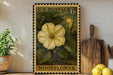 A framed U.S. postage stamp depicting a yellow flower, labeled "Moonflower", is displayed on a shelf next to a wooden cutting board and a potted plant.