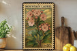 A framed vintage postage stamp depicting a pink flower plant, placed on a shelf next to a wooden cutting board and a potted plant.