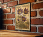 A framed illustration of a pasque flower is displayed on a brick wall, with a wooden table below it.