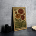 A metal stamp featuring a floral design with three large red flowers and two yellow flowers, placed on a dark surface next to some black camera lenses.