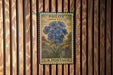 A vintage U.S. postage stamp featuring a blue flower illustration is displayed on a wooden wall.