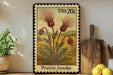 A framed postage stamp depicting a prairie smoke plant, with a wooden cutting board and a potted plant in the background.