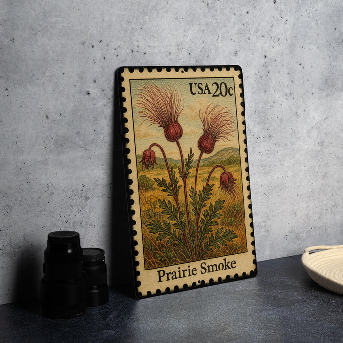 A framed postage stamp depicting a field of flowers, specifically Prairie Smoke, is displayed on a table.
