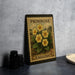 A metal sign with a floral design and text, likely a postage stamp, is displayed on a table next to some black camera lenses.