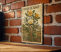 A framed illustration of a plant with yellow flowers is hanging on a brick wall.