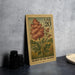 A wooden postcard with a floral design and text, sitting on a table next to some black camera lenses.