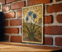a framed illustration of blue flowers on a brick wall, with the word "SCILLA" written above the flowers.