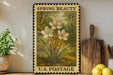 A framed vintage U.S. postage stamp depicting a floral scene, placed on a wooden surface next to a wooden cutting board and a potted plant.