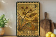 A vintage-style stamp depicting a witch hazel plant with yellow flowers, surrounded by autumn leaves, is displayed on a shelf next to a wooden cutting board and a potted plant.