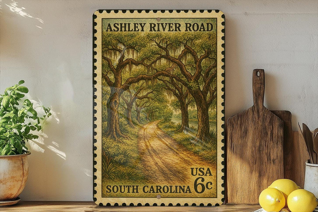 A framed vintage postage stamp depicting a scenic road through a lush, wooded area, with the text "ASHLEY RIVER ROAD" and "USA 6c SOUTH CAROLINA" printed on it. The stamp is displayed on a shelf next to a wooden cutting board and a potted plant.