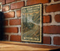 A vintage-style metal sign with a mountain landscape and a winding road, mounted on a brick wall.