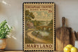 A framed illustration of a scenic countryside in Maryland, featuring a bridge, a house, and a river, is displayed on a shelf next to a wooden cutting board and a potted plant.